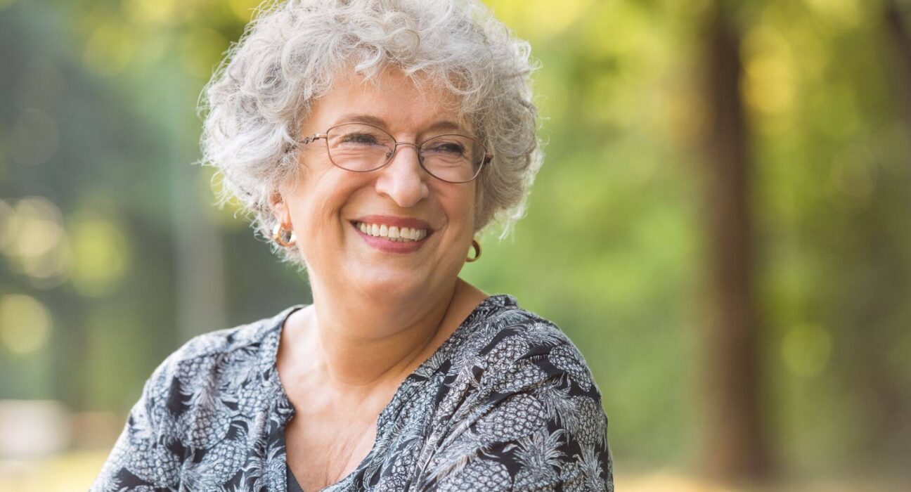 Uma mulher idosa sorrindo ao ar livre, usando óculos e uma blusa estampada, com um fundo desfocado de árvores e luz natural.