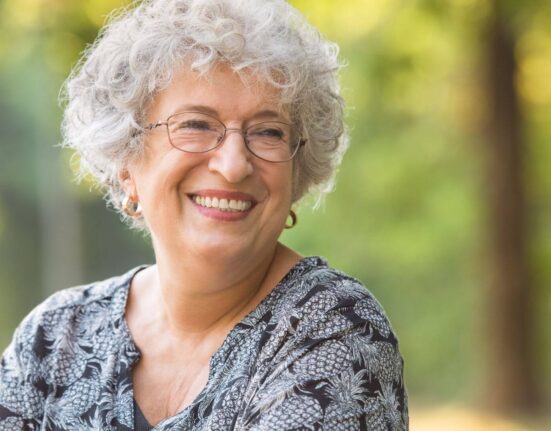 Uma mulher idosa sorrindo ao ar livre, usando óculos e uma blusa estampada, com um fundo desfocado de árvores e luz natural.